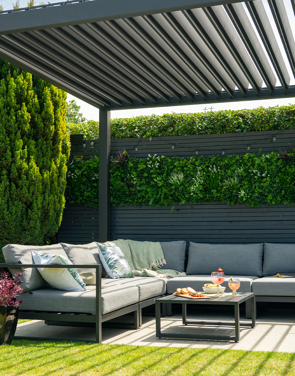 A landscape shot of our aluminium garden corner sofa set underneath a pergola, in a green garden.